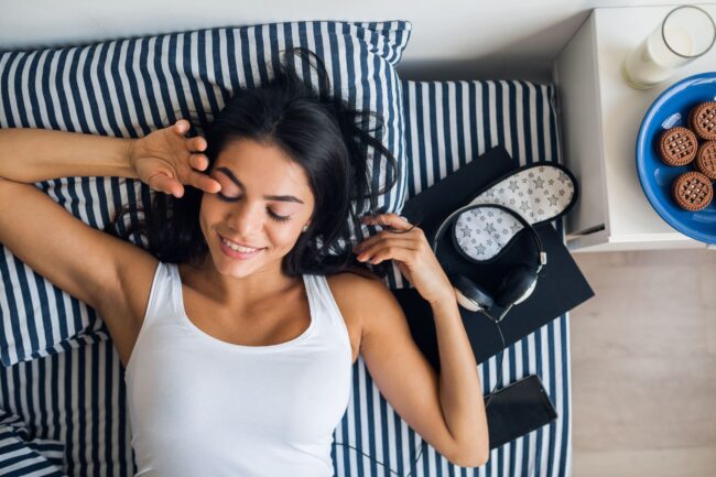 portrait de jeune femme seduisante couchee dans son lit en souriant vue d en haut se reveiller le matin paresseux loisirs dimanche detente petit dejeuner telephone casque livre masque de sommeil (1)